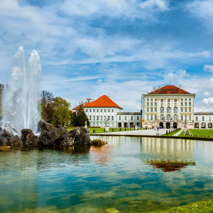 Im Vordergrund ein großer Springbrunnen. Im Hintergrund steht das Schloss Nymphenburg.