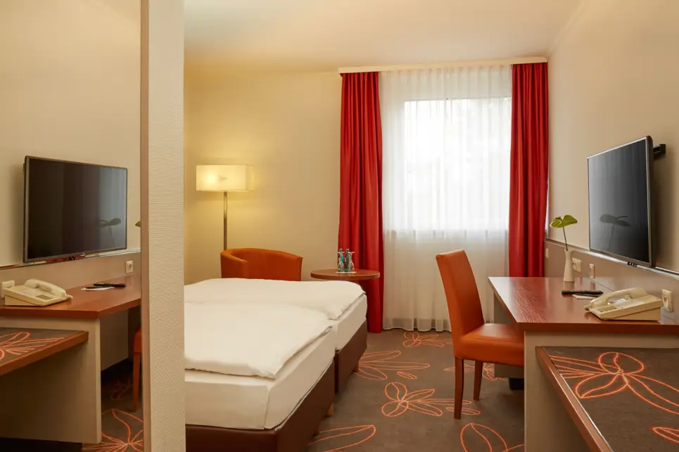 Blick ins Hotelzimmer mit einem Doppelbett auf der linken Seite und einem Schreibtisch auf der rechten Seite. Am Eingangsbereich hängt ein großer Spiegel.