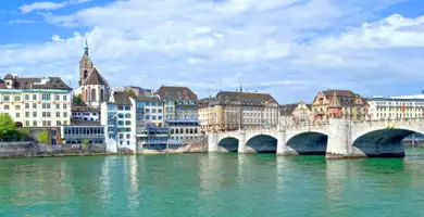 Blick über den blauen Rhein auf die Basler Rheinpromenade mit einer Brücke.