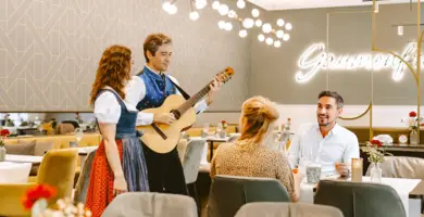 Two guests are sitting in the restaurant of the HYPERION Hotel Salzburg. A man is playing the guitar at their table and a singer is standing next to him.