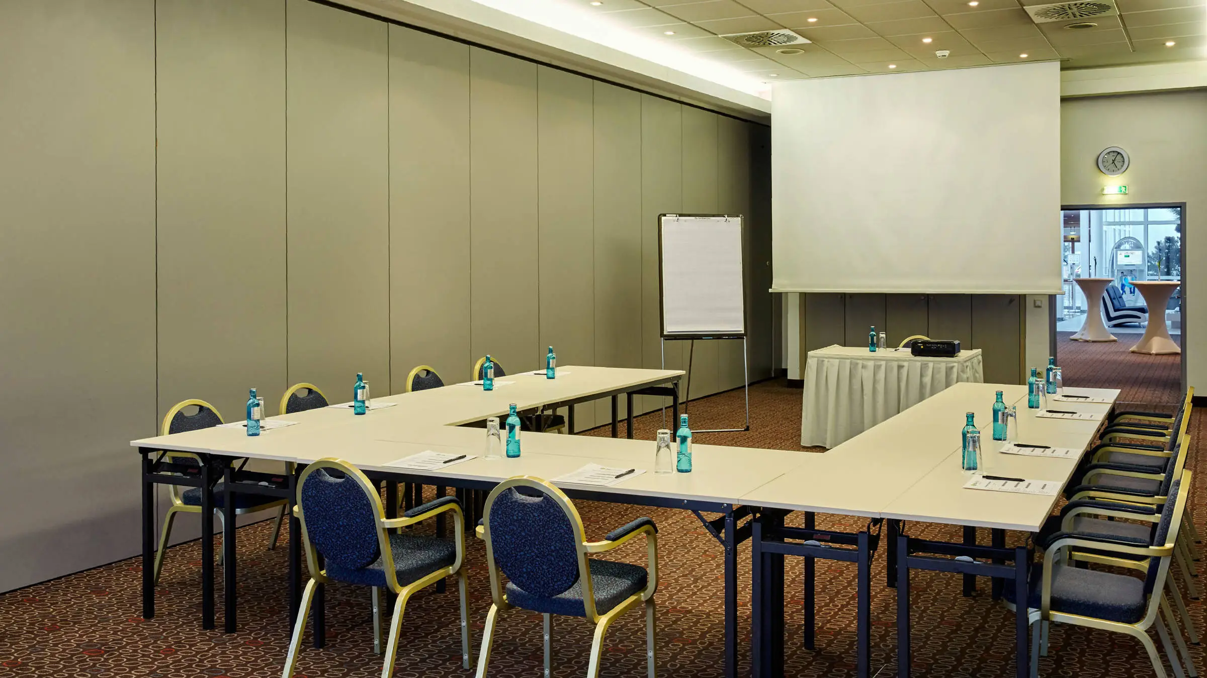 Tagungsraum mit UForm Bestuhlung. In der Mitte steht ein Rednertisch mit einer großen Leinwand.
