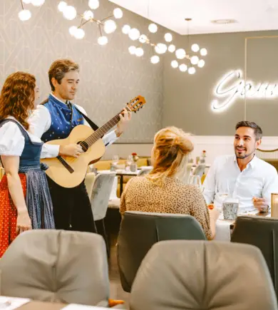 Two guests are sitting in the restaurant of the HYPERION Hotel Salzburg. A man is playing the guitar at their table and a singer is standing next to him.