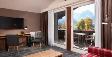 Hotelzimmer mit einem Schreibtisch und einem Fernseher an der Wand. Auf der rechten Seite sind bodentiefe Fenster mit Blick auf den Balkon und Berge.