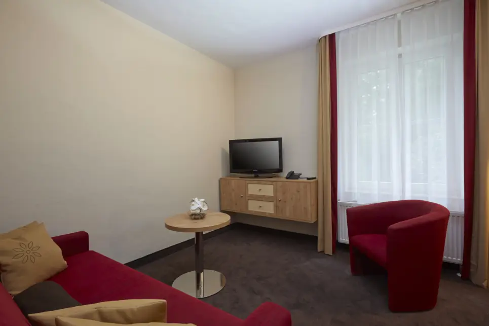 Zona de estar con un sofá y un sillón. Hay una pequeña cómoda con un televisor en la pared.