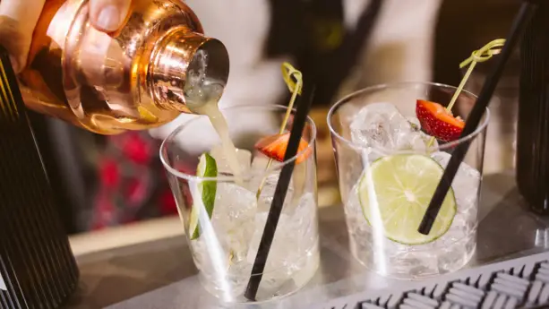 2 Gläser mit Eiswürfel, schwarzen Strohhalmen, Limettenscheiben und jeweils einem Stück Erdbeere. In das linke Glas wird ein Getränk aus einem Cocktail-Shaker eingegossen.