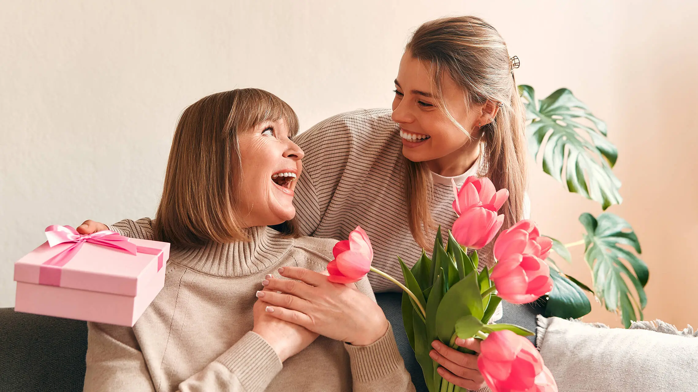 Two women look at each other with a smile. One woman is holding flowers and a gift.