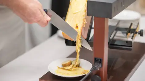 Ein Koch schneidet mit einem Messer geschmolzenen Käse aus einem Käserad für Raclette.