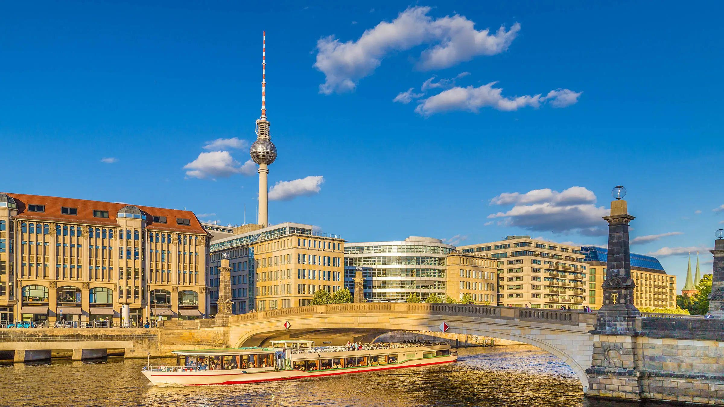 Blick über die Spree zum Berliner Fernsehturm. Auf der Spress fährt ein Ausflugsboot unter einer Brücke.