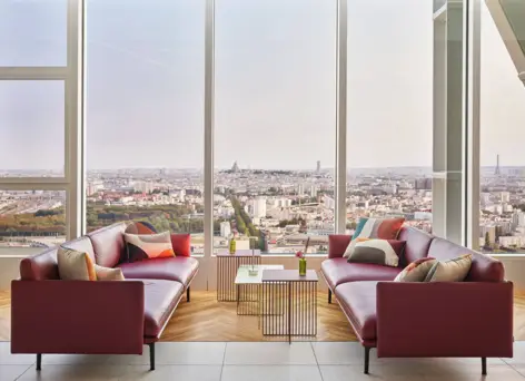 Two burgundy sofas with tables in front of large windows overlooking Paris.