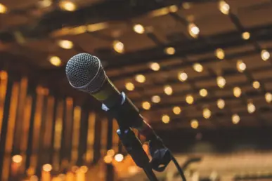 Microphone in a stand on a stage in front of empty rows of chairs. Bright light bulbs on the ceiling.