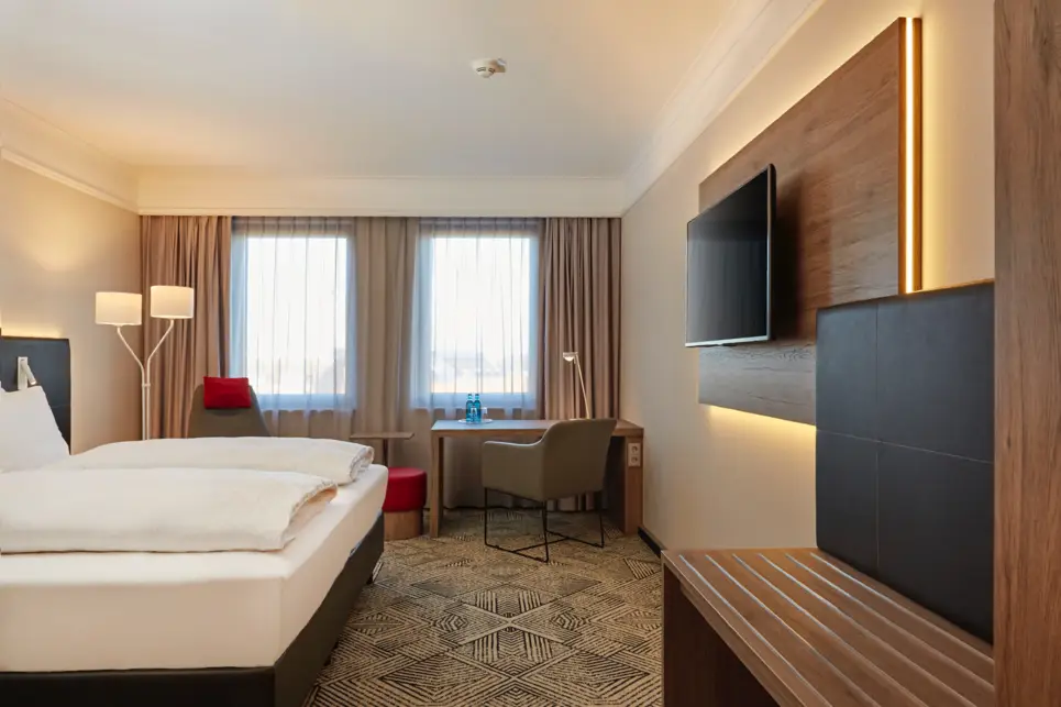 Hotelzimmer mit einem Doppelbett auf der linken Seite und einem Fernseher an der Wand auf der rechten Seite. Dahinter steht am Fenster ein Tisch mit einem Stuhl.