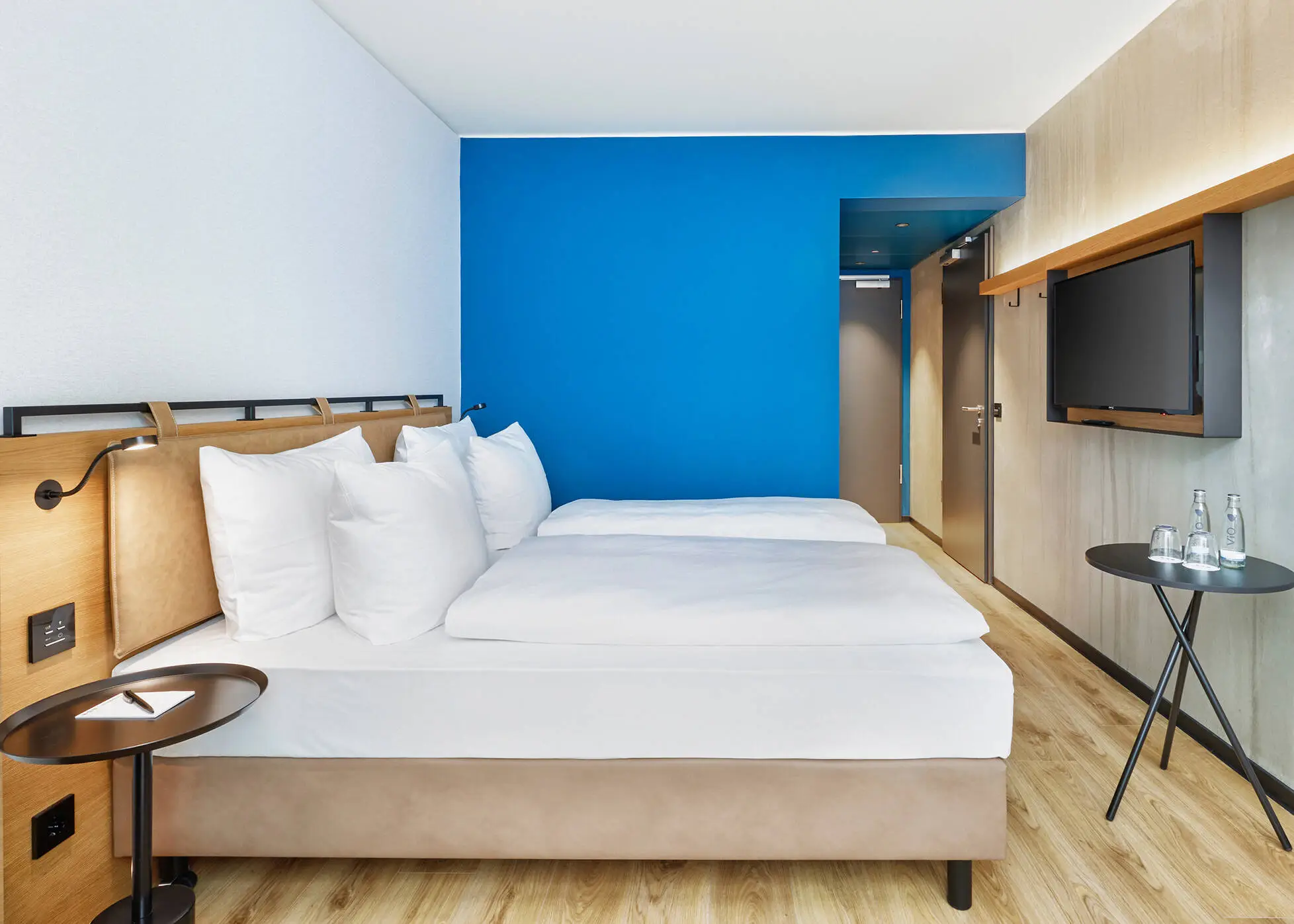 Hotelzimmer mit einem Doppelbett in dunkelbraunen und blauen Farben. Vor dem Bett hängt ein Fernseher.