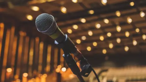 Mikrofon in einem Ständer auf einer Bühne vor leeren Stuhlreihen. Leuchtende Glühbirnen an der Decke.