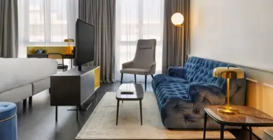 Hotelzimmer mit einer großen, dunkelblauen Couch auf der rechten Seite. Auf der linken Seite steht ein kleiner Fernsehschrank mit einem Fernseher darauf.