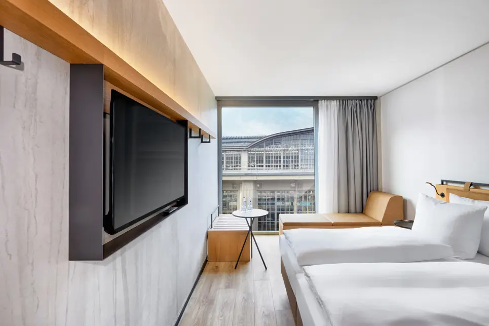 Sehr helles Hotelzimmer mit einem Doppelbett und einem Fernseher an der Wand. Durch das Fenster sieht man den Hauptbahnhof.
