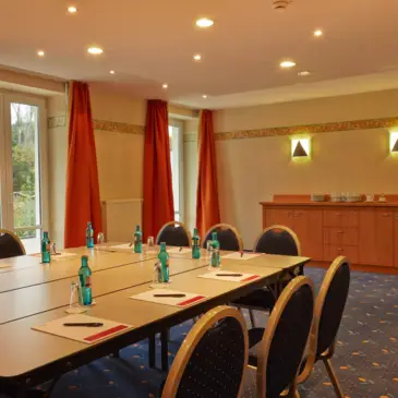 Großer Konferenztisch vor großen Fenstern. An der Wand dahinter steht eine kleine Kommode mit Kaffeeservice