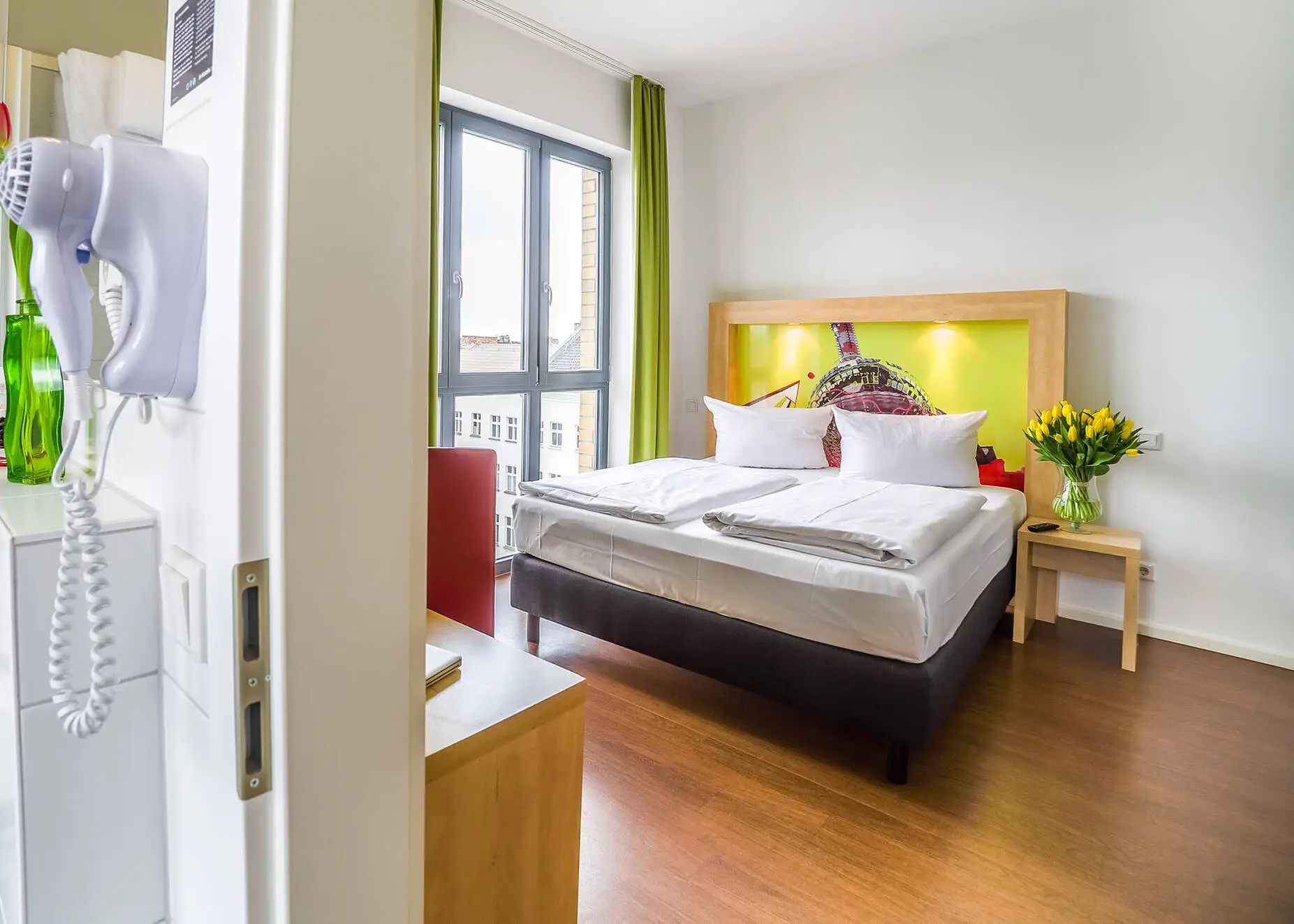Blick vom Badezimmer in das Hotelzimmer mit Doppelbett. Das Badezimmer hat neben dem Waschbecken einen Föhn hängen.