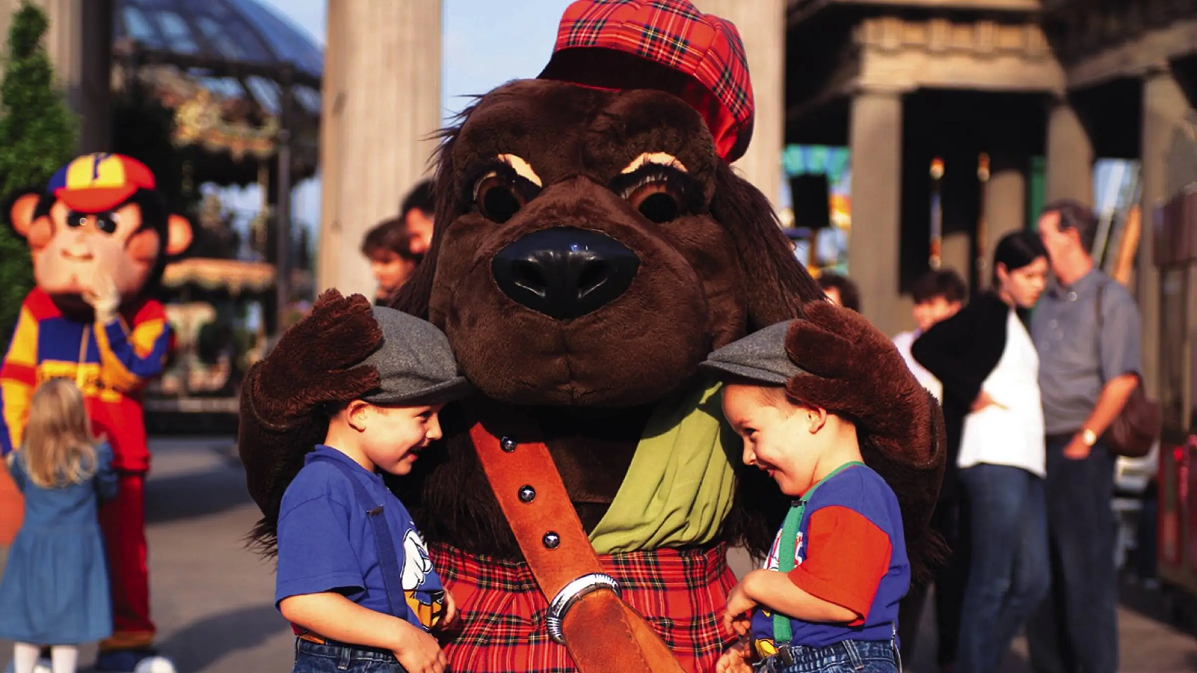 Two small children stand happily in front of a dog mascot. The two children look at each other, the mascot has both paws on the children's heads.