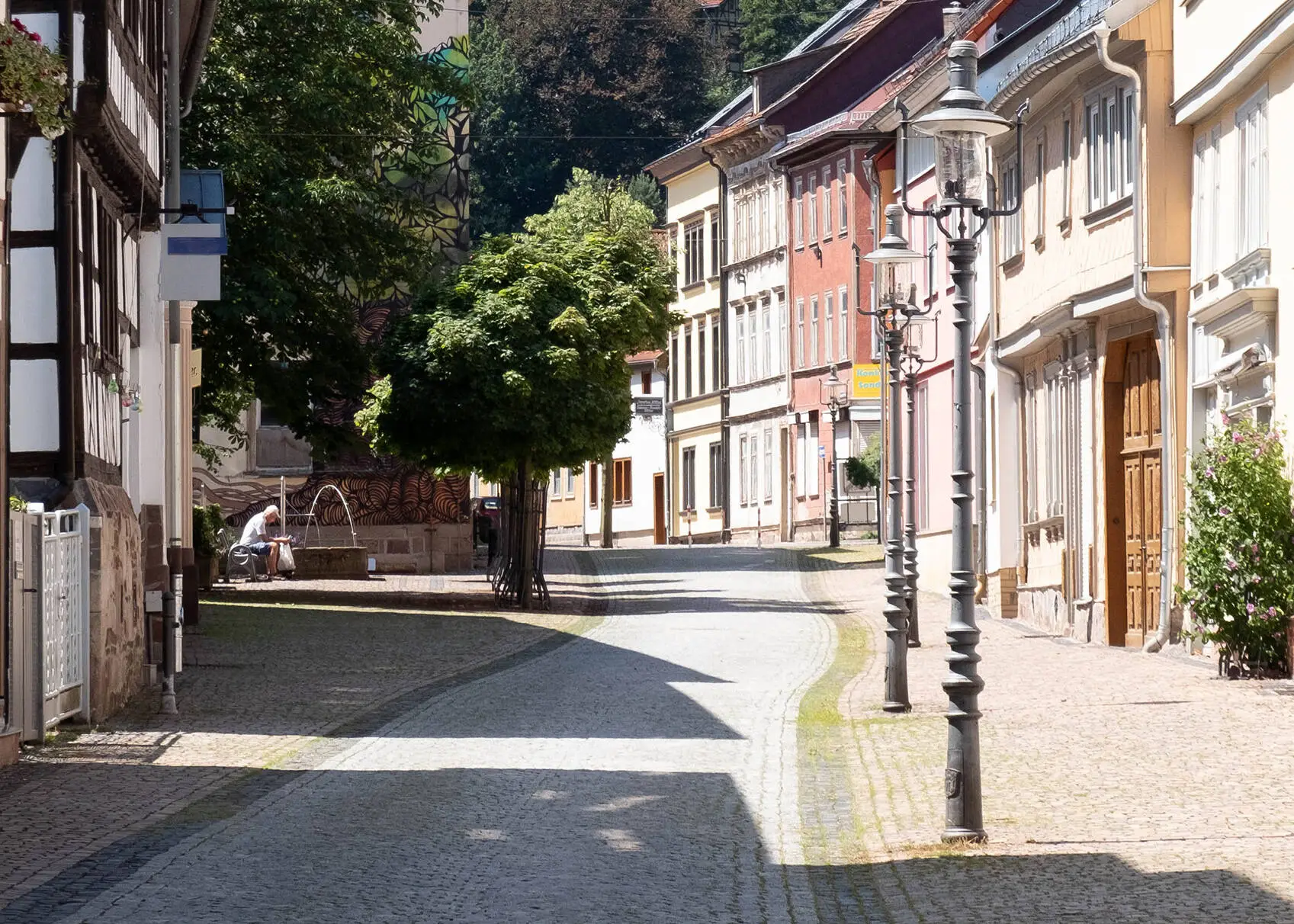 Idyllic street in the summery old town of Friedrichroda with half-timbered houses and lanterns lining the roadside. In the background, trees and a man sitting on a bench next to a fountain.