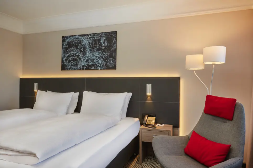 Auf der linken Seite steht ein Doppelbett über dem ein Bild mit mathematischen Gleichungen. Auf der rechten Seite steht ein grauroter Sessel.