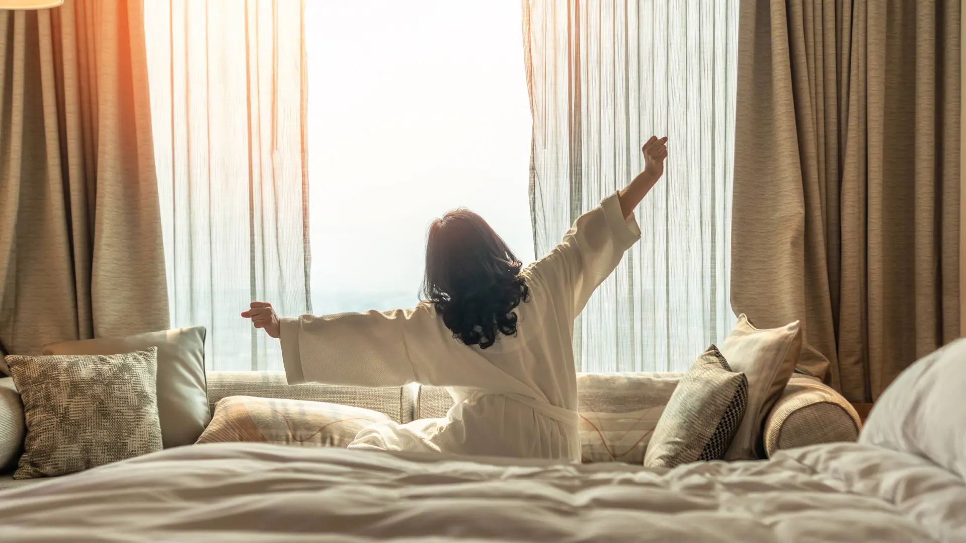 A woman in a bathrobe sits on the bed and stretches.