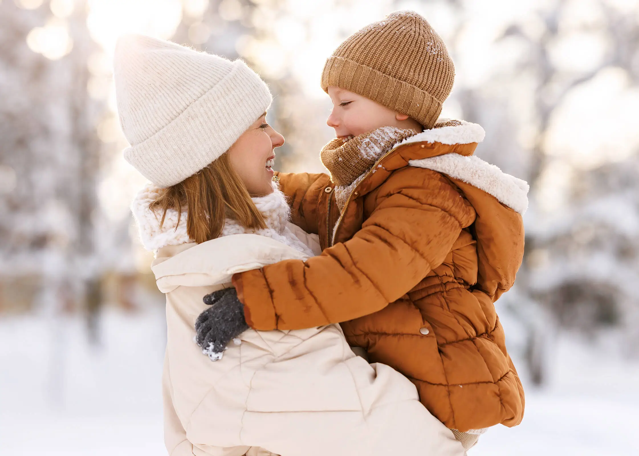 Eine Frau  mit weißer Jacke und Mütze hält ein Kind in brauner Jacke und Mütze. Im Hintergrund liegt viel Schnee.