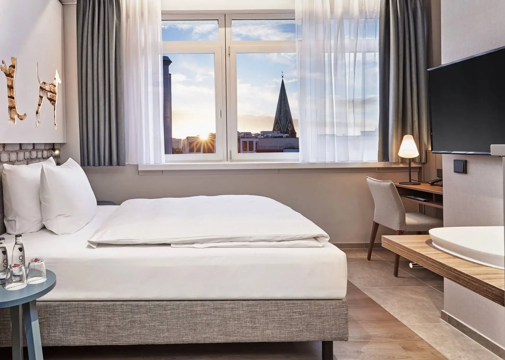 Chambre d'hôtel avec un lit et un bureau devant. Au premier plan, sur la gauche, se trouve une armoire avec une machine à café et sur la droite, une télévision est suspendue.