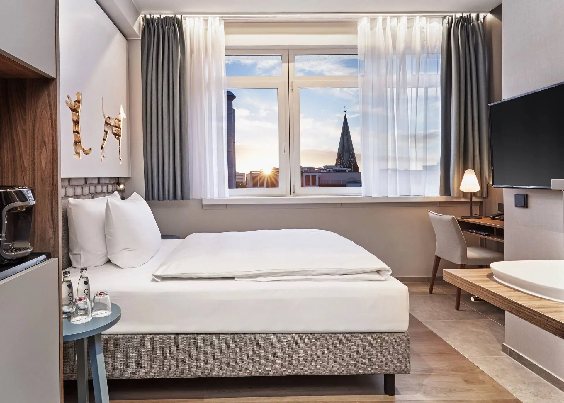 Hotelzimmer mit einem Bett und einem Schreibtisch davor. Im Vordergrund steht auf der linken Seite ein Schrank mit einer Kaffeemaschine und auf der rechten Seite hängt ein Fernseher.