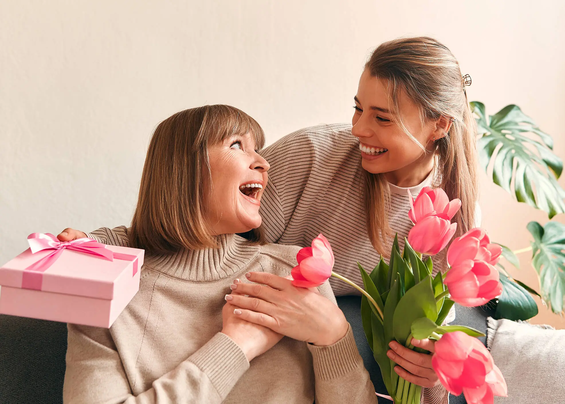 Two women look at each other with a smile. One woman is holding flowers and a gift.