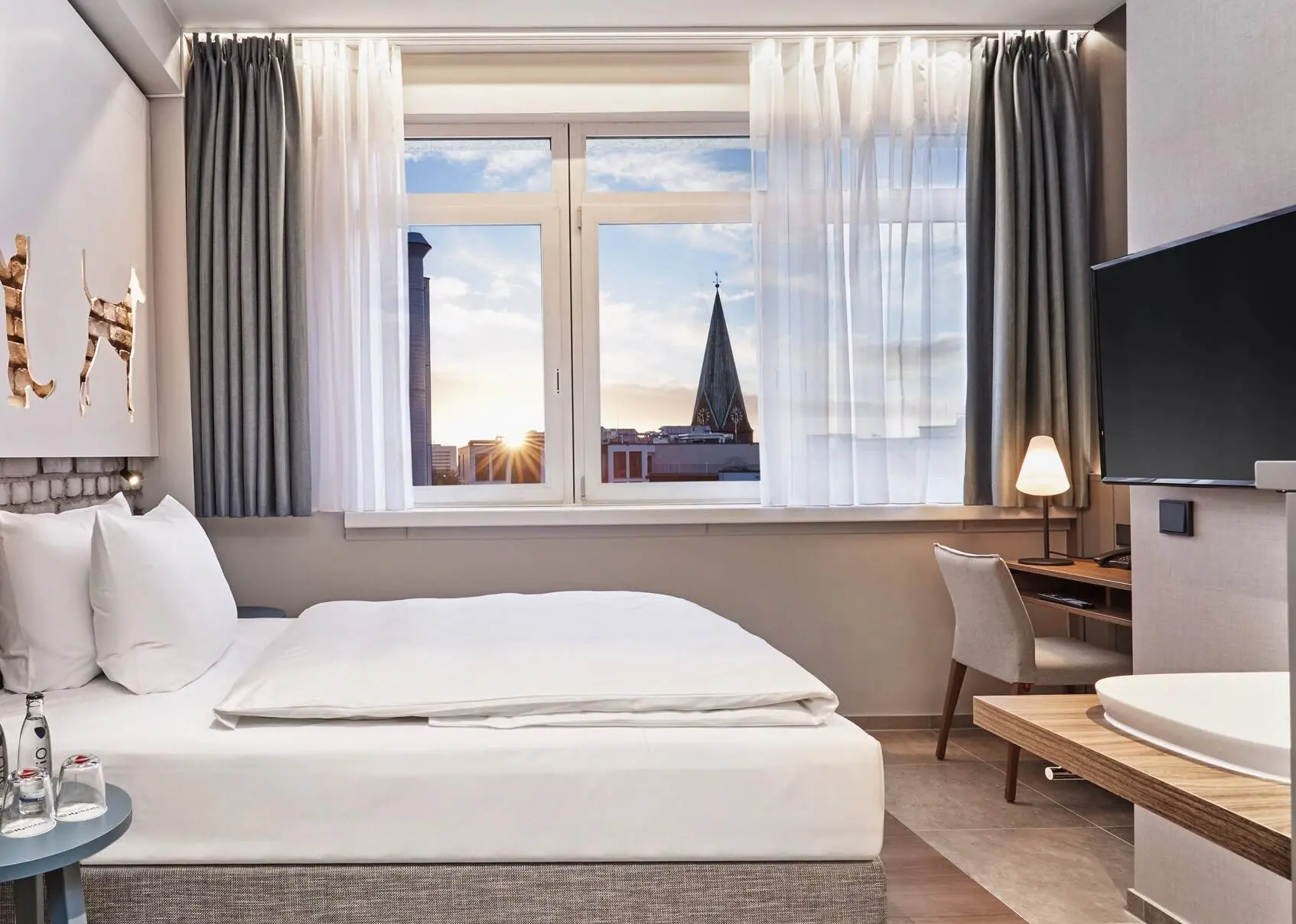 Habitación de hotel con una cama y un escritorio delante. En primer plano a la izquierda hay un armario con una cafetera y a la derecha cuelga un televisor.