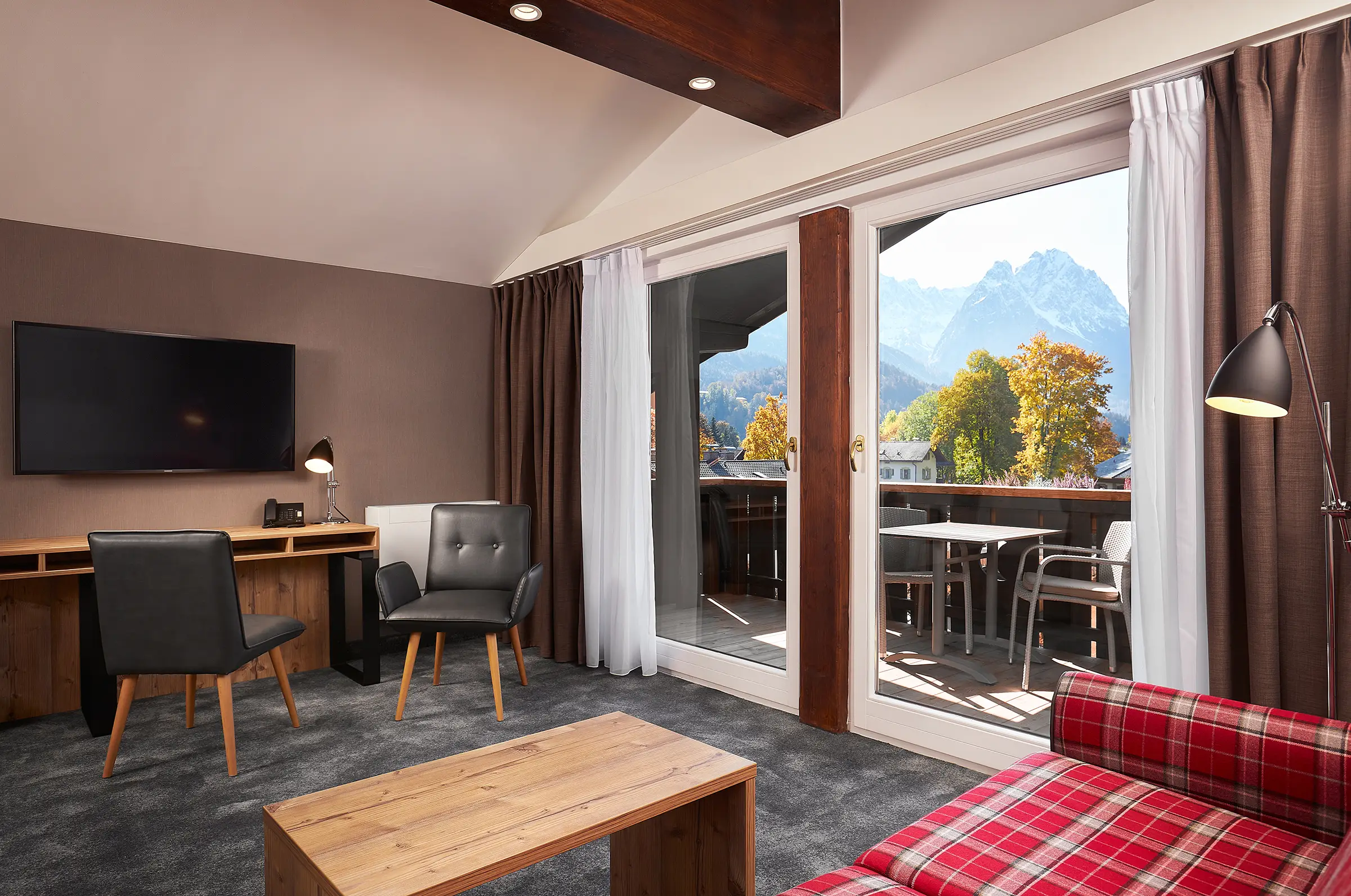 Blick vom Sofa aus dem Zimmer auf den Balkon und das Bergpanorama. Auf der linken Seite steht ein Schreibtisch mit 2 Stühlen vor einem hängenden TV an der Wand.