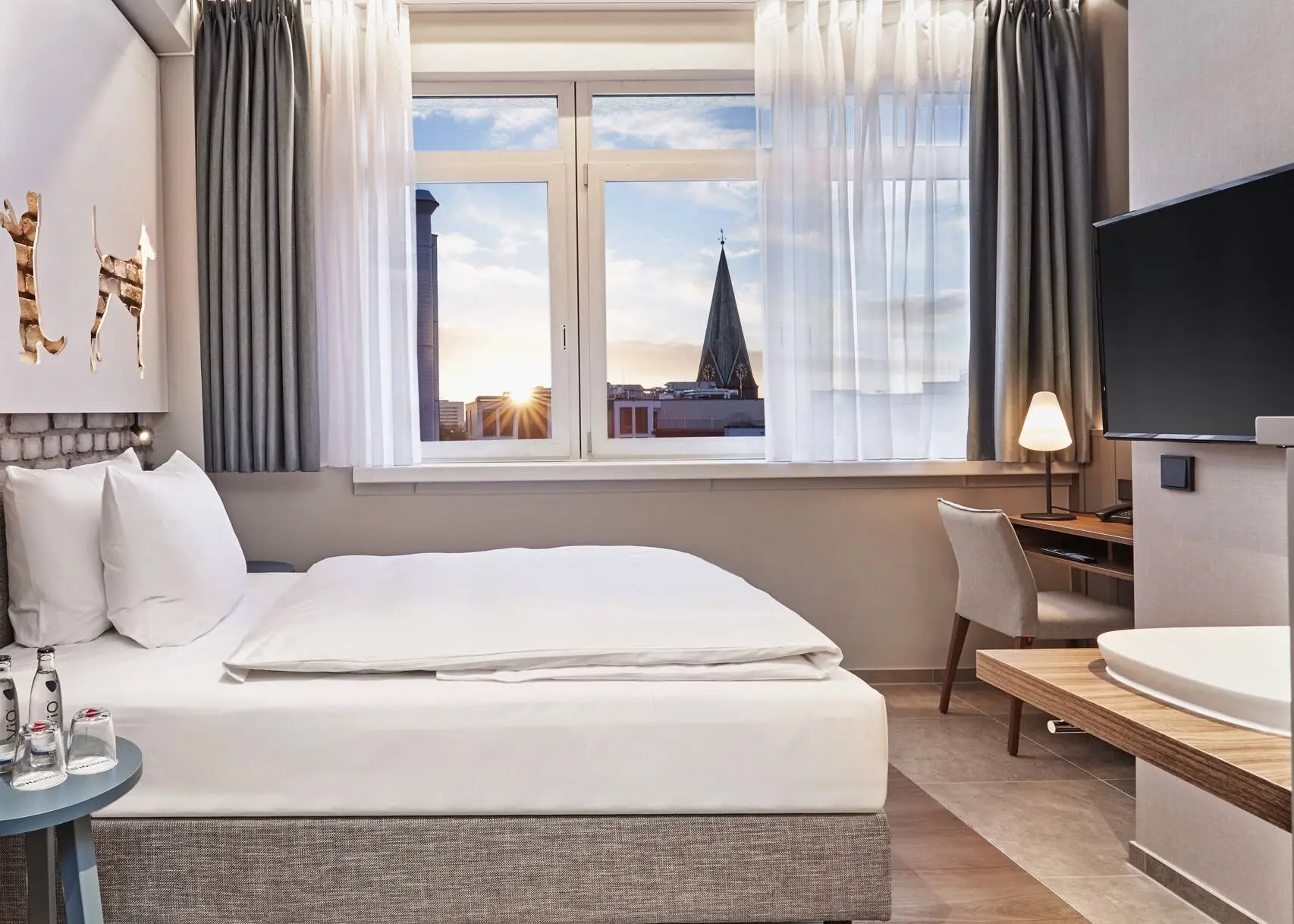 Hotel room with a bed and a desk in front of it. In the foreground on the left is a cupboard with a coffee machine and on the right hangs a television.