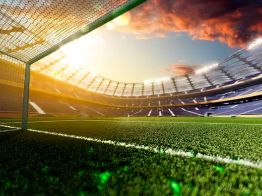 View across the pitch from the goal at sunset in a football stadium. The setting sun and clouds can be seen in the sky.